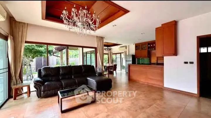 Spacious living room with elegant chandelier and open kitchen layout, featuring large windows and garden view.