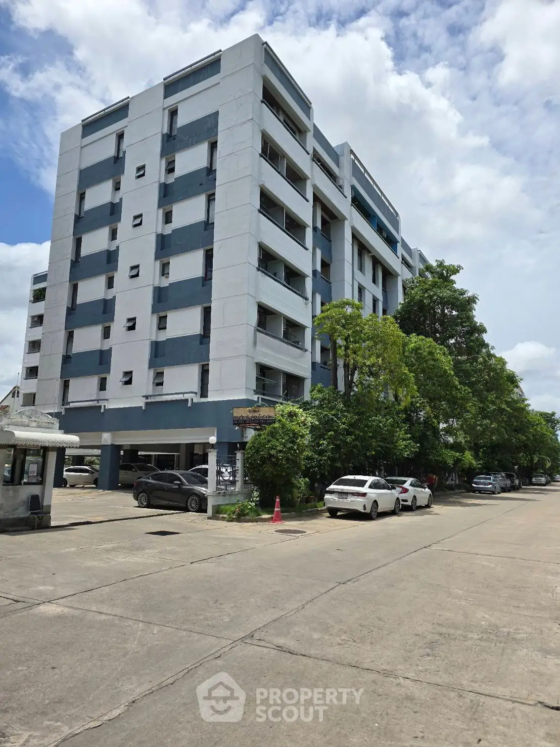 Modern multi-story residential building with parking and lush greenery under a clear blue sky.