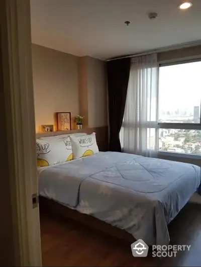 Sunlit bedroom with panoramic city views, featuring a large bed with decorative pillows, floor-to-ceiling windows, and warm wooden flooring.