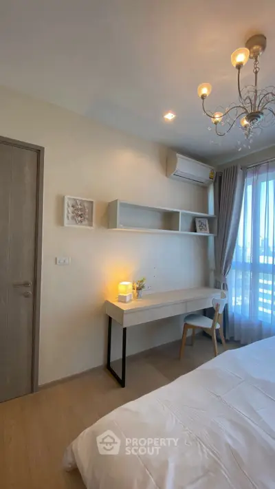 Modern bedroom with elegant chandelier, cozy workspace, and natural light from large window.