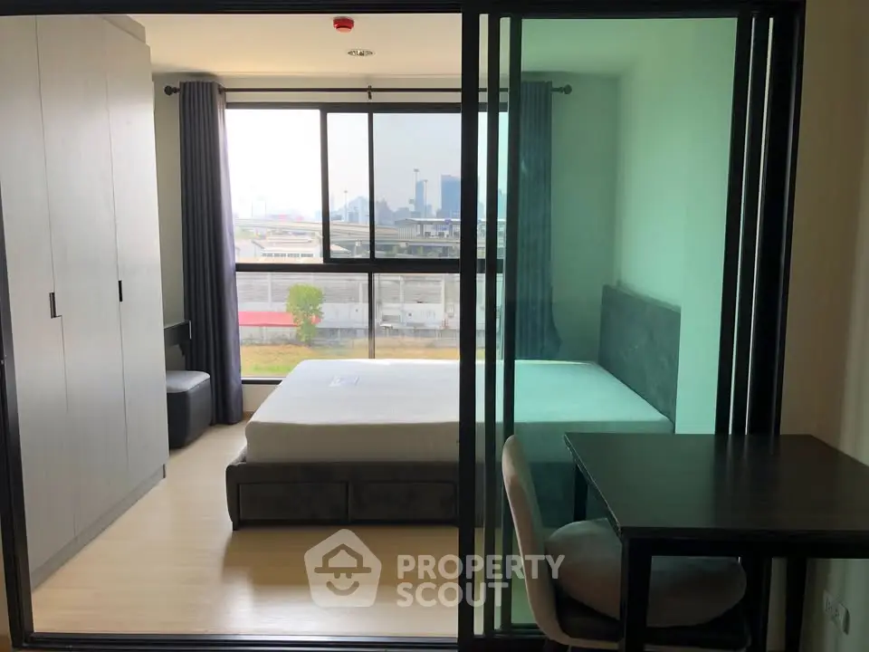 Modern bedroom with large window and city view, featuring a bed, wardrobe, and desk.