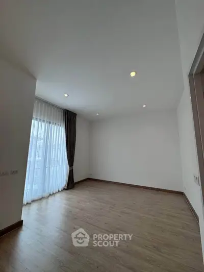 Spacious empty room with wooden flooring and large window with curtains.