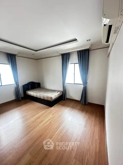 Spacious bedroom with wooden flooring and natural light, featuring a cozy bed and elegant curtains.