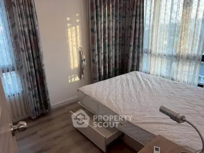 Cozy bedroom with natural light and floral curtains, featuring a comfortable bed and wooden flooring.