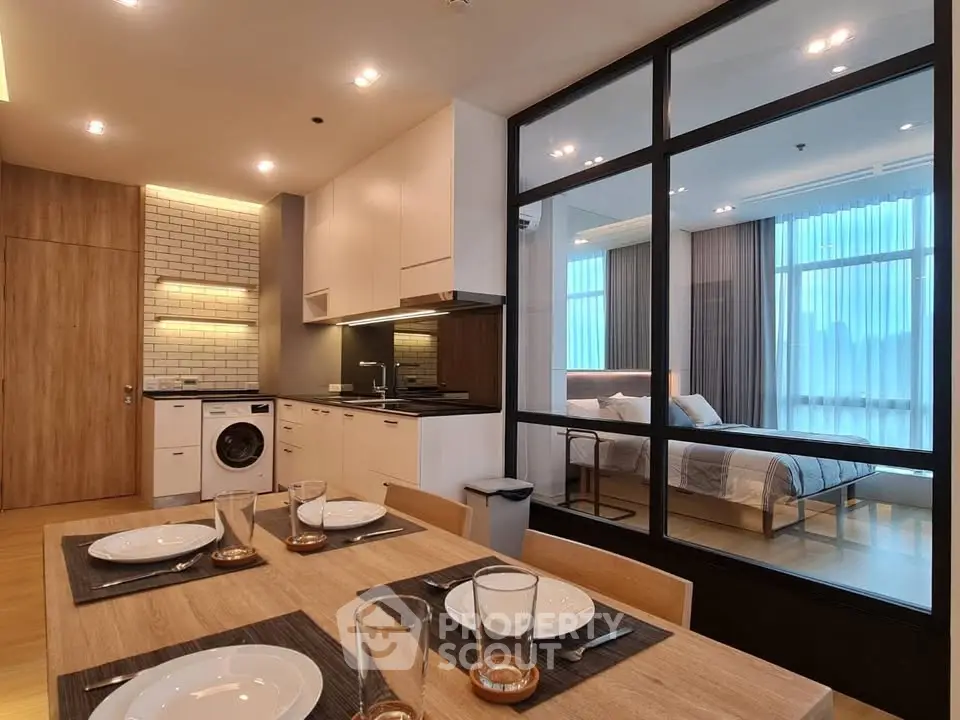 Modern open-plan kitchen and dining area with glass partition to bedroom, featuring sleek design and natural light.