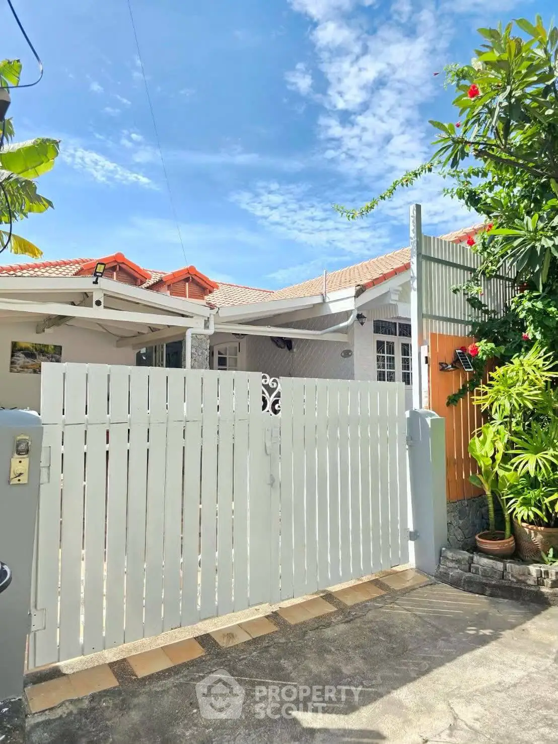 Charming single-story house with gated entrance and lush greenery under a clear blue sky.