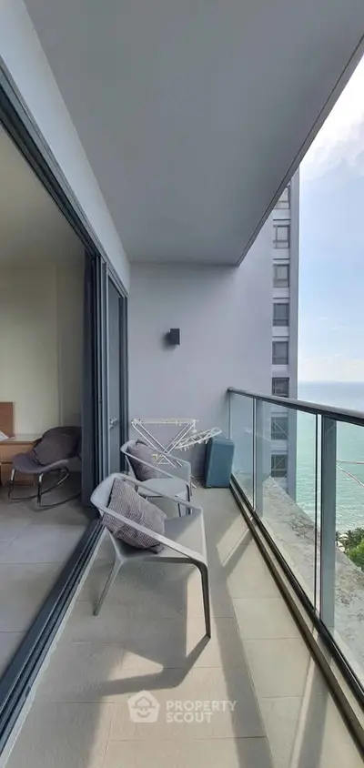 Modern balcony with ocean view, glass railing, and cozy seating in a high-rise apartment.