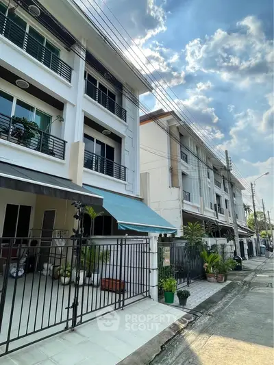 Modern townhouse exterior with gated entrance and lush greenery under a vibrant sky.