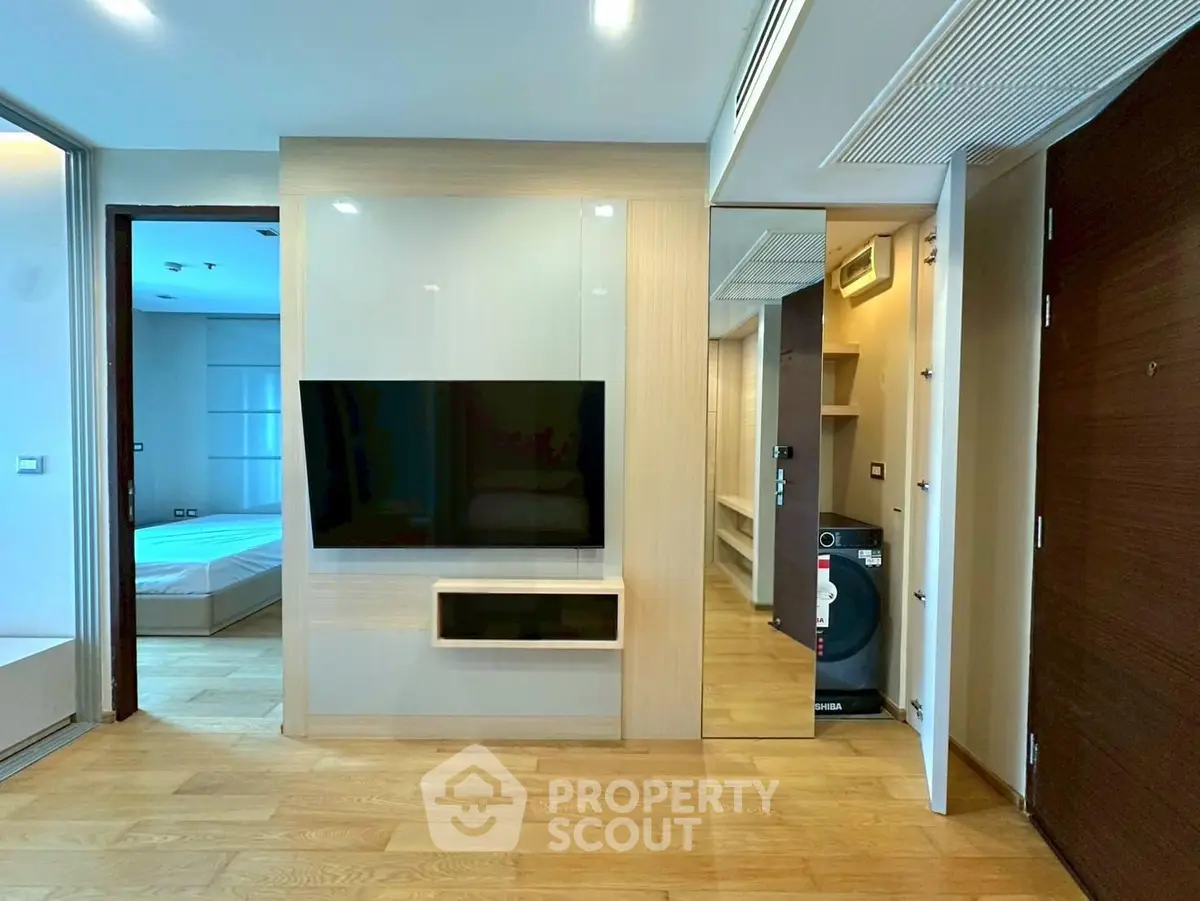 Modern apartment interior with sleek wooden flooring and built-in TV unit, adjacent to a cozy bedroom.
