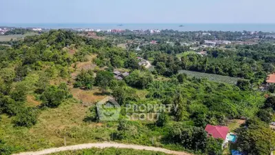 Expansive aerial view of a lush green plot of land with potential for development, surrounded by trees and with a distant ocean view, ideal for a luxury estate or resort.