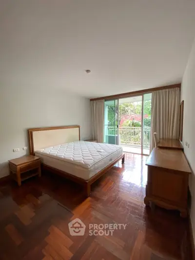 Spacious bedroom with hardwood floors and large window overlooking greenery, featuring a comfortable bed and wooden furniture.
