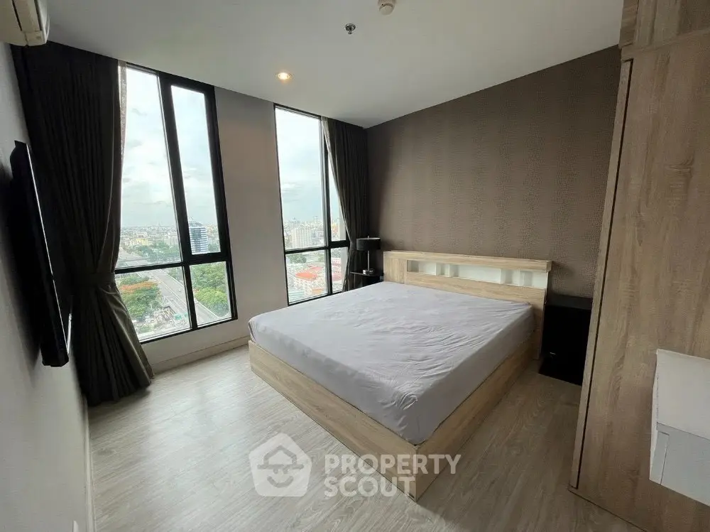 Modern bedroom with large windows and city view, featuring sleek wooden furniture and neutral decor.
