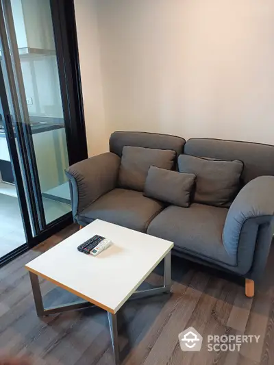 Cozy modern living room with comfortable grey sofa and minimalist white coffee table, featuring sleek hardwood flooring and ample natural light from large glass door.