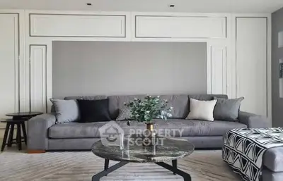 Elegant living room with a plush grey sofa, chic patterned ottoman, and a sleek glass coffee table, set against a backdrop of sophisticated white paneling.