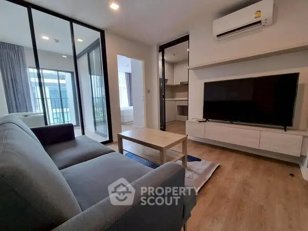 Modern living room with sleek furniture and large TV, adjacent to a kitchen area.