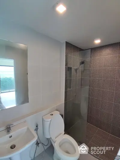 Modern bathroom with sleek mosaic tiles, well-lit with natural light, featuring a glass shower area, toilet, and basin.