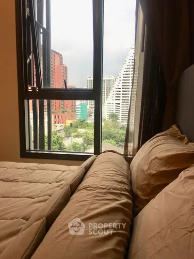 Cozy bedroom with city view through large window, featuring modern high-rise buildings.