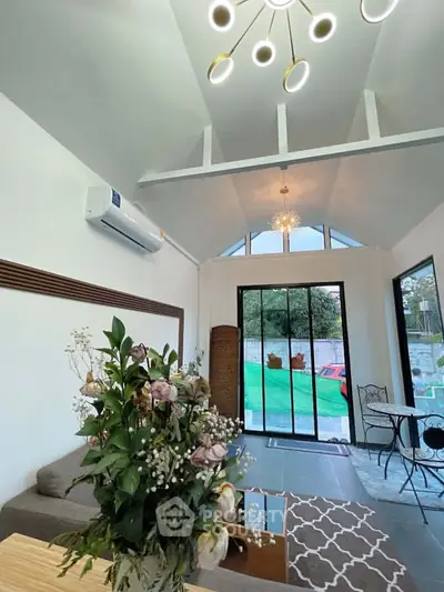 Modern living room with high ceiling and large windows opening to a garden view, featuring elegant decor and lighting.
