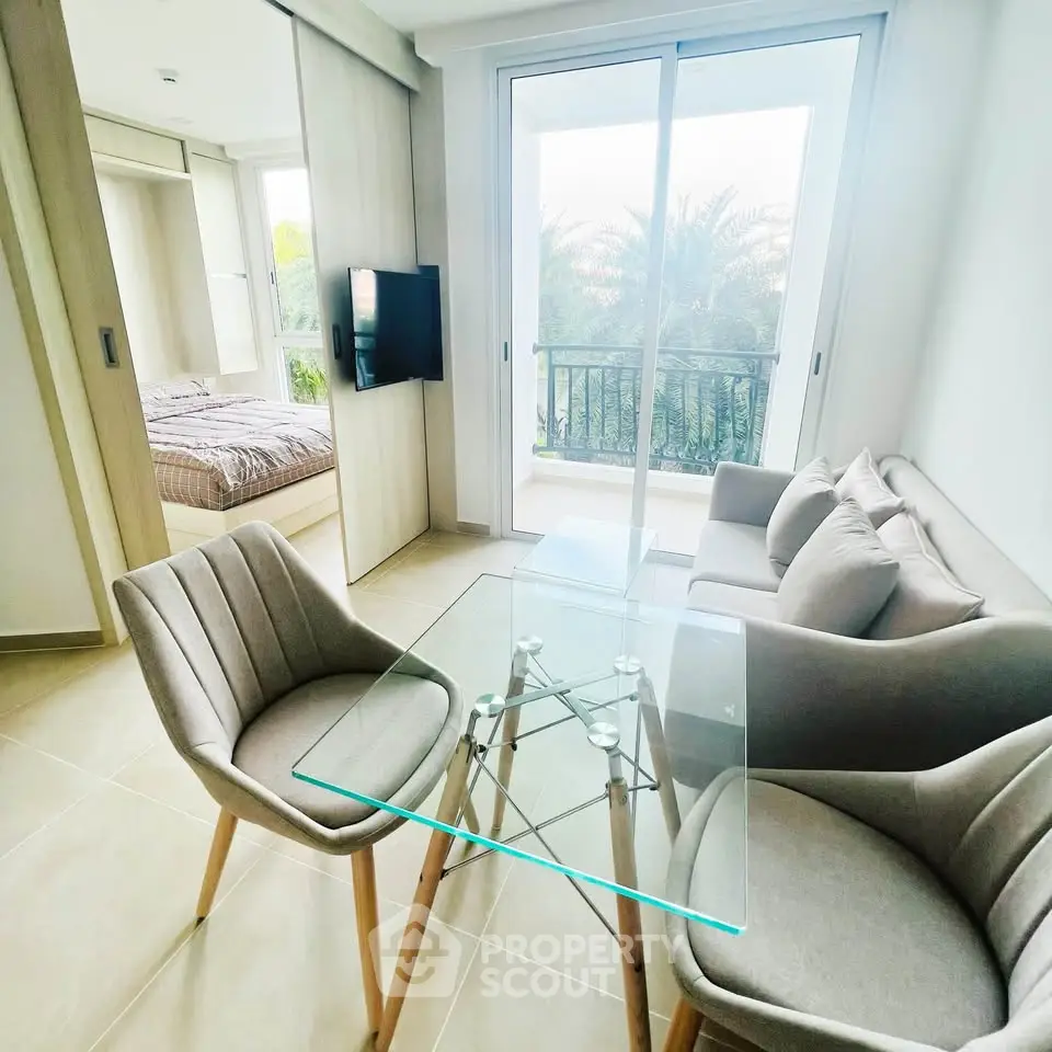 Modern living room with glass table and cozy seating, adjacent to a bedroom with natural light.