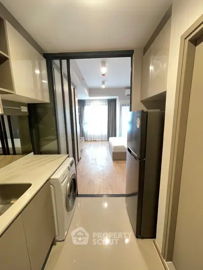 Modern apartment kitchen with washing machine and fridge leading to a bright living area.
