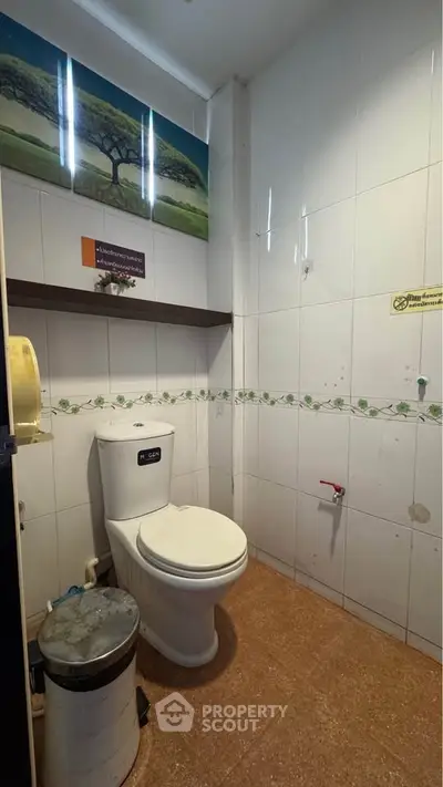 Compact bathroom with white tiles and modern toilet, featuring wall art and a small trash bin.