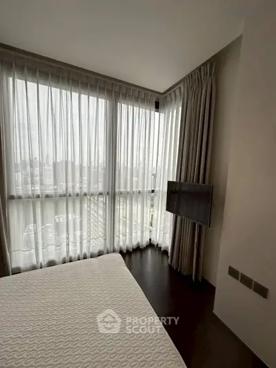 Modern bedroom with floor-to-ceiling windows and city view, featuring a wall-mounted TV and elegant curtains.