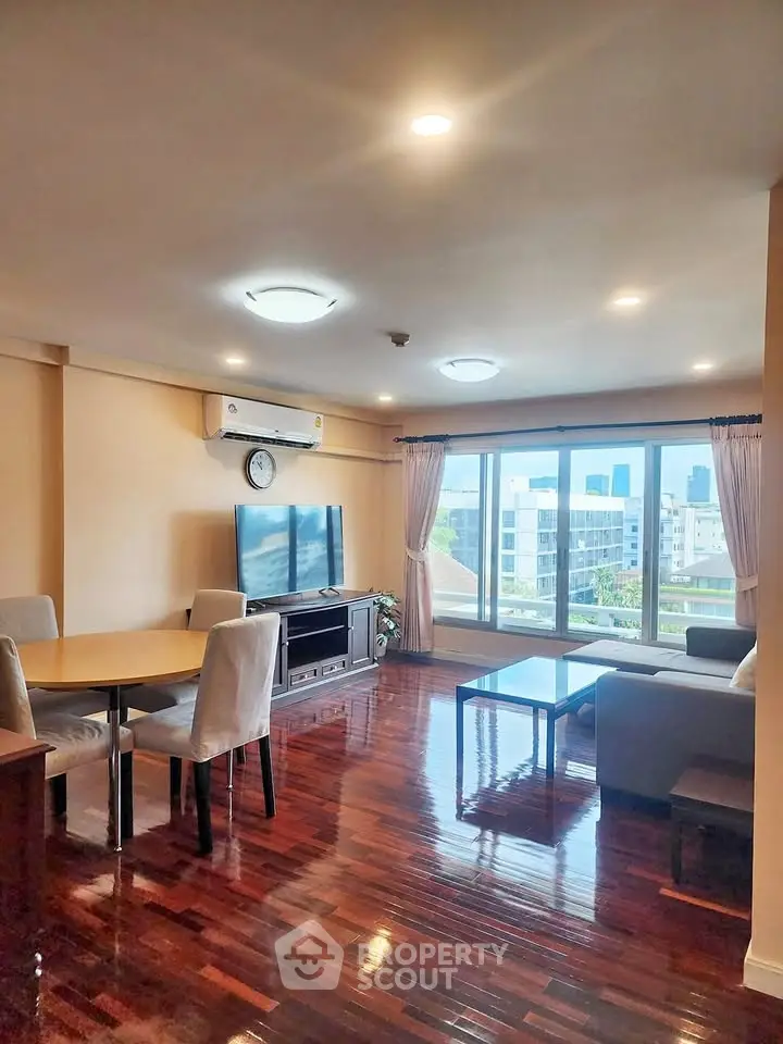 Spacious living room with hardwood floors and city view, featuring modern furniture and large windows.