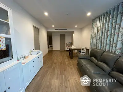 Spacious living room with elegant hardwood flooring, plush leather sofa, and modern lighting, leading to an open-plan kitchen area.