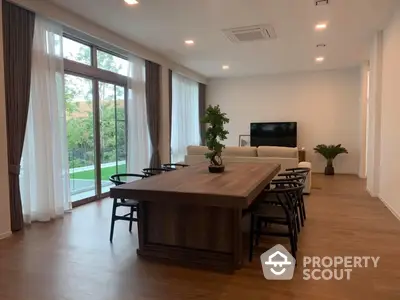 Spacious and modern living room with large windows offering ample natural light, elegant wooden flooring, and direct access to a serene outdoor view, perfect for family gatherings.