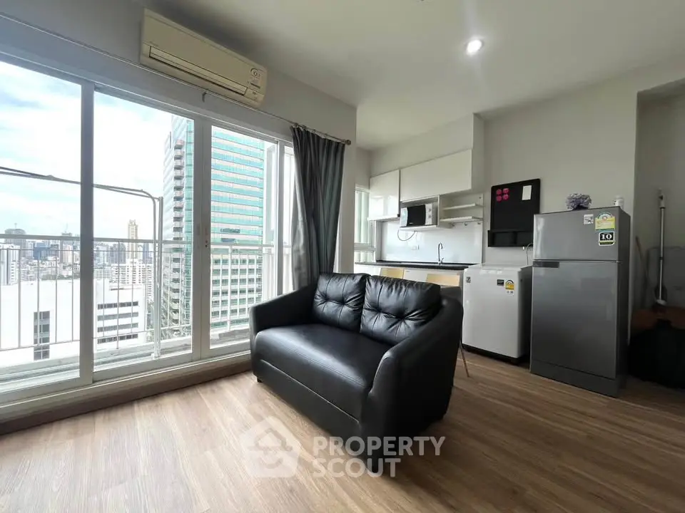 Modern living room with city view, featuring sleek black sofa and compact kitchen area.