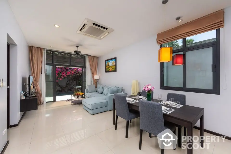 Spacious and modern living room with sleek dining set, comfortable sofa, and vibrant hanging lights, leading to a cozy balcony with pink flowers.