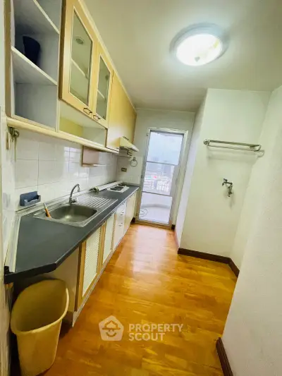 Compact kitchen with wooden flooring and closed layout, featuring a sink and stove, leading to a balcony.