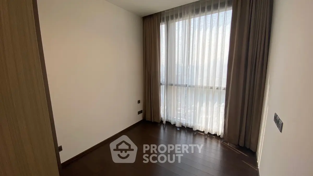 Spacious bedroom with large window and elegant curtains, featuring dark wood flooring.