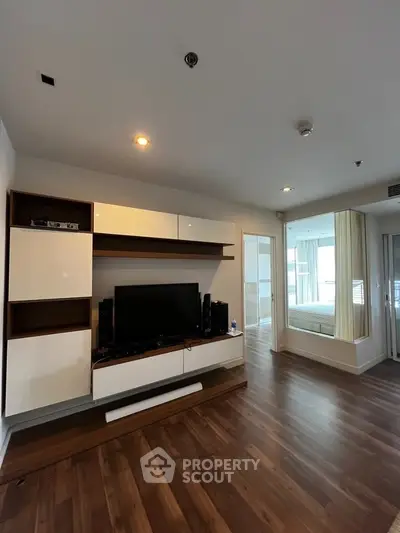 Modern living room with sleek wooden flooring and built-in entertainment unit, leading to a bright bedroom.