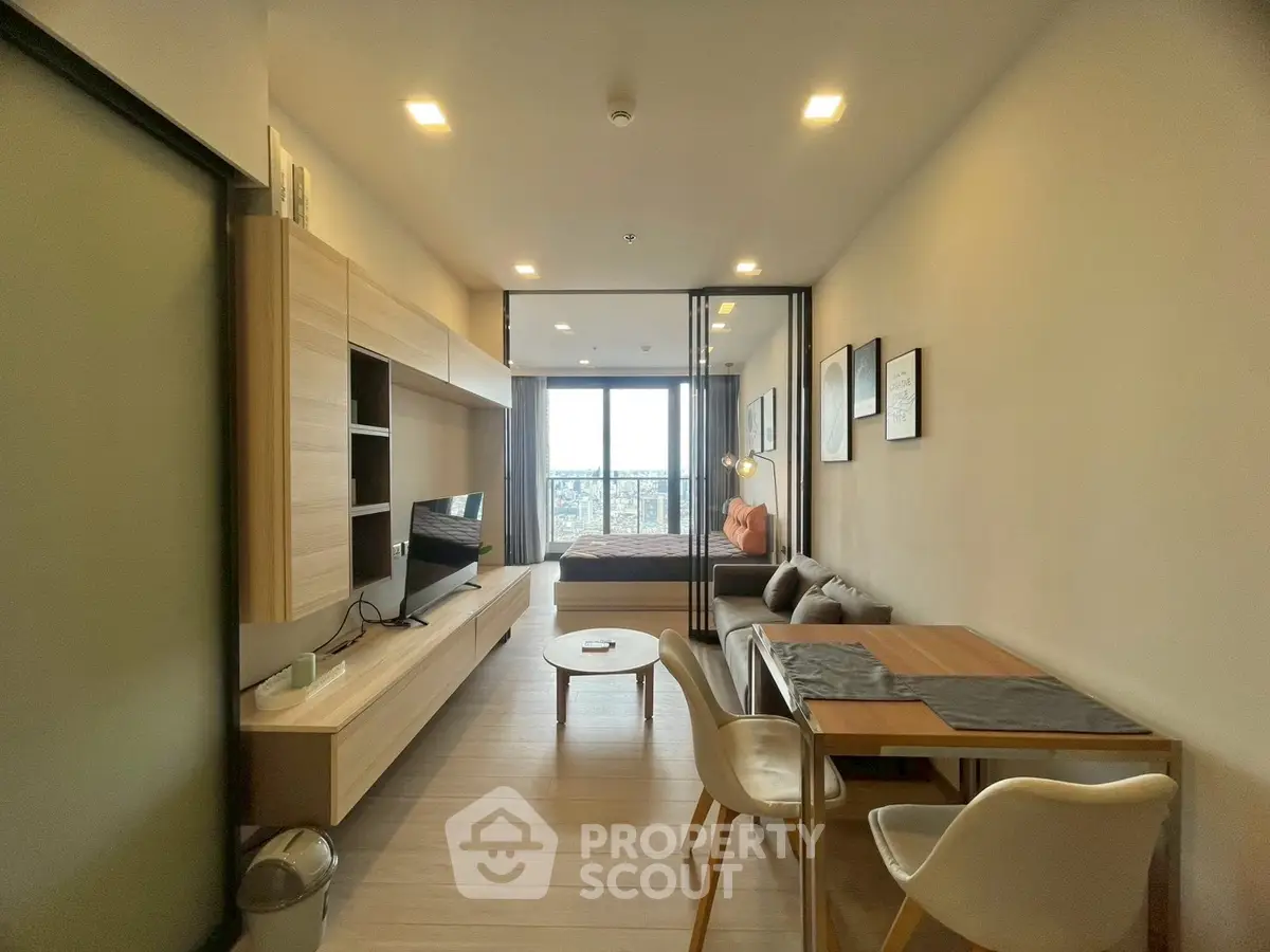 Modern open-plan living room with dining area and bedroom, featuring sleek furniture and large windows.