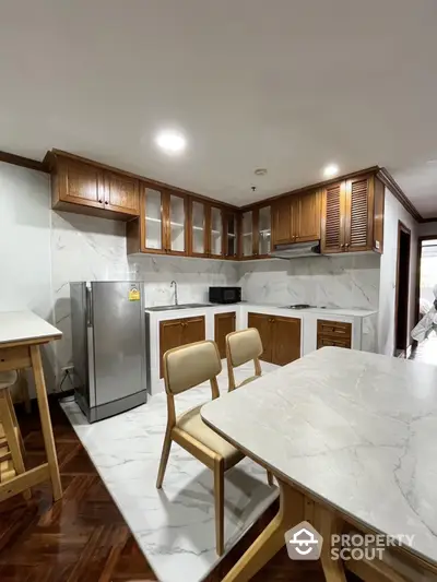 Modern kitchen with marble countertops and wooden cabinets, featuring a sleek refrigerator and dining area.