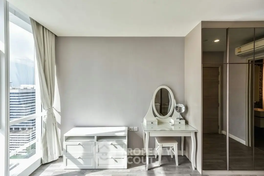 Modern bedroom with sleek furniture and city view, featuring a stylish vanity and ample natural light.
