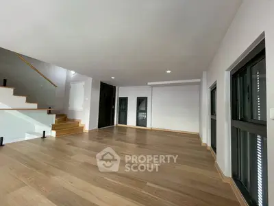 Spacious modern living room with wooden flooring and staircase, ample natural light from large windows.