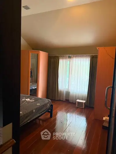 Cozy bedroom with wooden flooring and large window with curtains, featuring a comfortable bed and wardrobe.