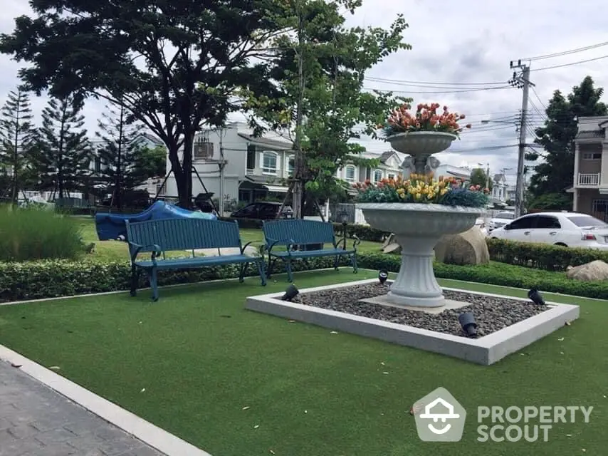 Charming community garden with vibrant green artificial turf, elegant benches, and a classic fountain centerpiece, surrounded by lush landscaping and residential backdrop.