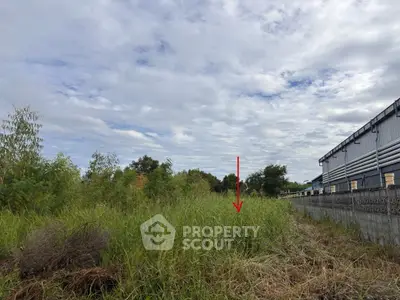 Vacant land with overgrown grass and industrial building nearby, ideal for development.