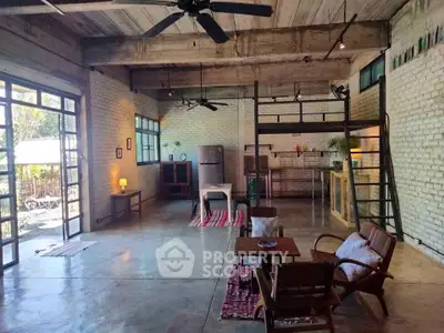Spacious industrial-style living room with open kitchen and loft area, featuring exposed brick and large windows.