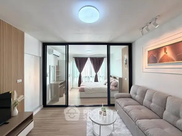 Modern living room with glass partition leading to a cozy bedroom, featuring stylish decor and natural light.