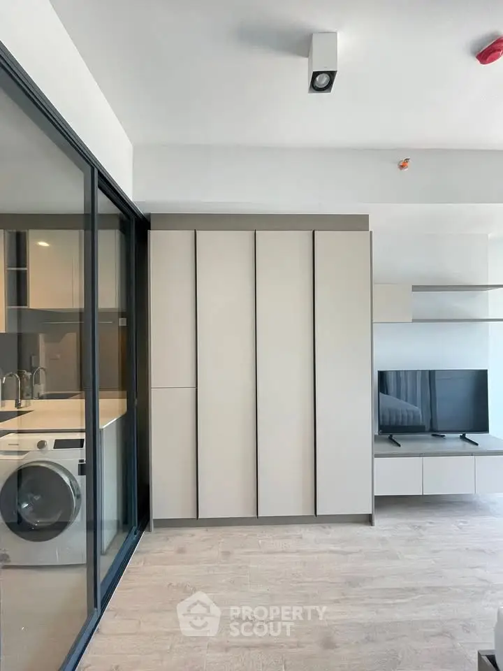 Modern living room with sleek cabinetry and built-in TV unit, adjacent to a compact kitchen with washing machine.