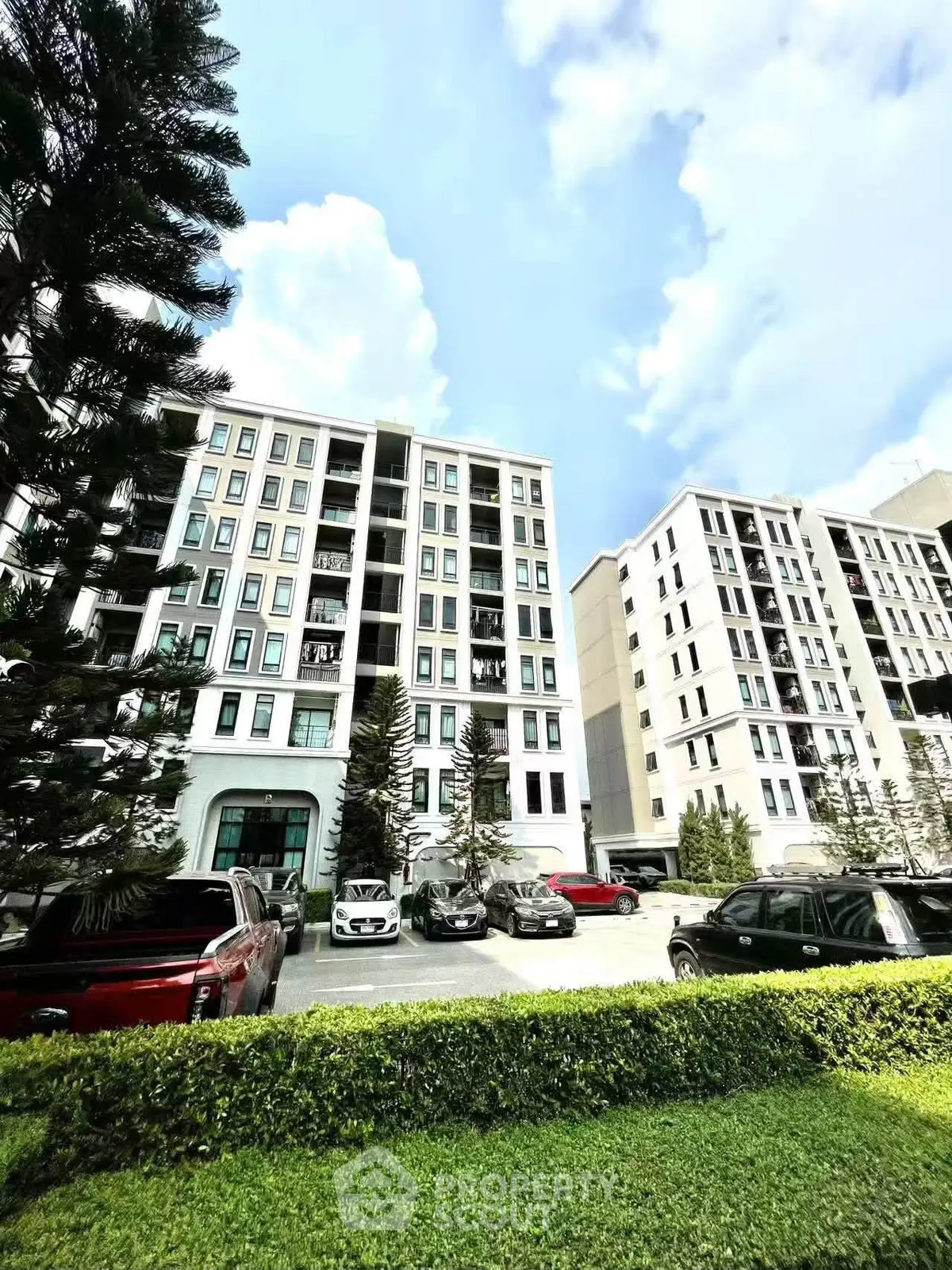 Modern apartment building with lush greenery and parking area under a clear blue sky.