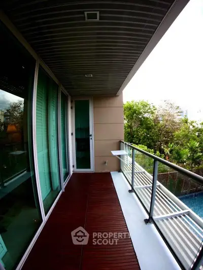 Spacious balcony with wooden flooring and glass railing overlooking lush greenery.