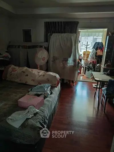 Cozy bedroom with wooden flooring and natural light, featuring a bed and cluttered storage area.