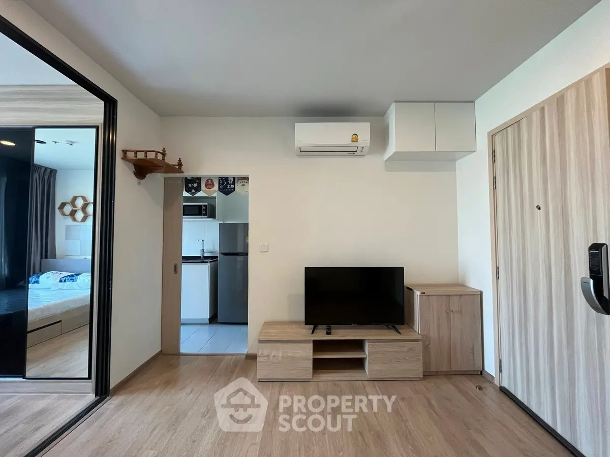 Modern living room with wooden flooring, TV unit, and view into a compact kitchen and bedroom.