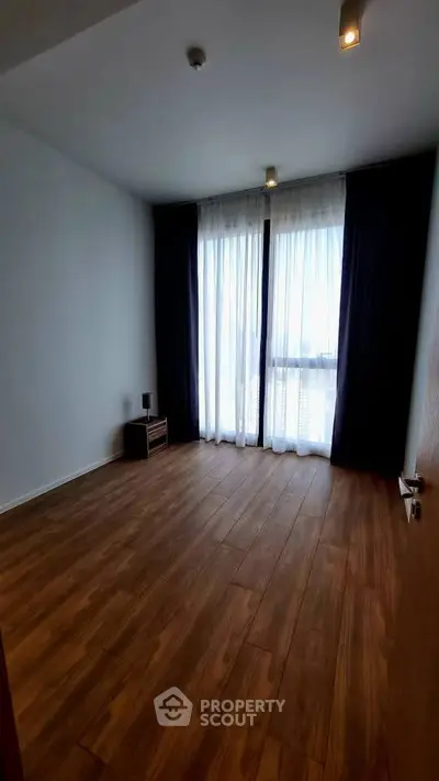Spacious empty room with wooden flooring and large windows with curtains.