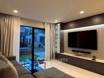 Modern living room with large TV, sleek furniture, and view of outdoor area through glass doors.